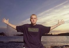 a man with his arms wide open sitting on a rock by the water at sunset