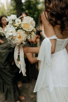 a woman in a white dress holding a bouquet of flowers with other people behind her