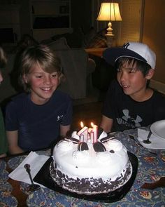 three children are sitting at a table with a birthday cake in front of them that has lit candles on it