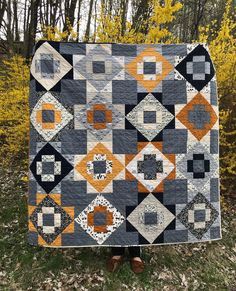 a person holding up a quilt in front of some trees and grass with yellow leaves