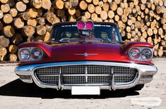 an old red car is parked in front of some logs and wood stacked up behind it