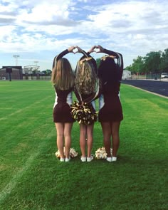 three girls standing in the middle of a field making a heart shape with their hands