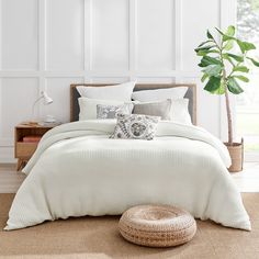 a bed with white sheets and pillows in a room next to a potted plant