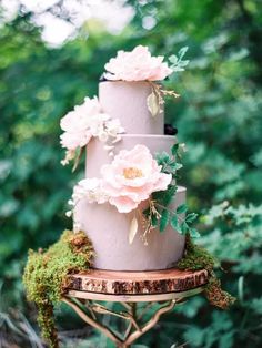 a three tiered cake with pink flowers on top is surrounded by moss and trees