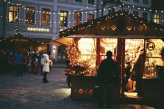 people are walking around an outdoor market with christmas lights on the buildings in the background