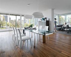 a glass dining table surrounded by white chairs in a living room with hardwood floors and large windows