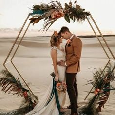 a bride and groom standing in the sand with their arms around each other under a gold geometric frame