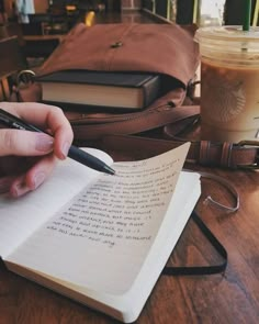 a person is writing in a notebook with a pen and coffee on the table next to them