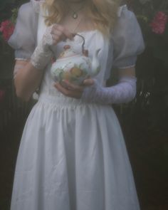 a woman in a white dress holding a tea pot and wearing gloves with flowers behind her