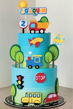 a three tiered cake with cars, trucks and trees on the top is decorated in blue icing