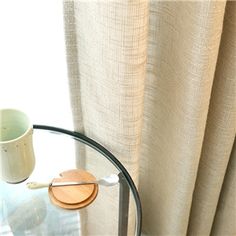 a round glass table with a wooden object on it next to a white cup and curtain