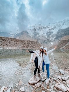 two women standing on rocks in front of a lake