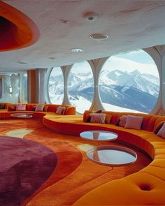 an orange couch sitting inside of a living room next to large round windows with snow covered mountains in the background