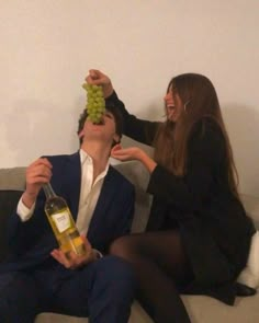 two people sitting on a couch and one is holding a bottle of wine while the other holds a bunch of grapes