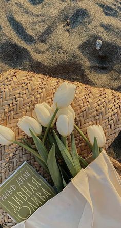 some white tulips are sitting on the sand near a basket with a book