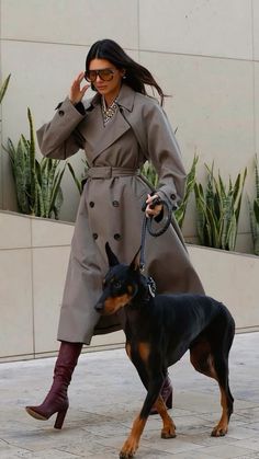 a woman in a trench coat and boots walking her dog on a leash while talking on the phone