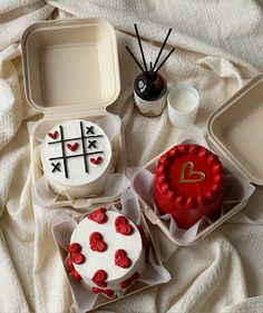 three cakes in plastic containers on a bed with white sheets and one has a red heart