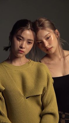 two young women standing next to each other in front of a dark background, one wearing a green sweater