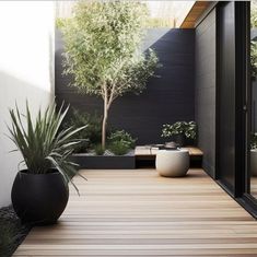 a wooden deck with potted plants and trees in the back yard, next to a black wall