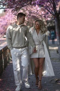 a man and woman walking down a sidewalk under trees with purple flowers in the background