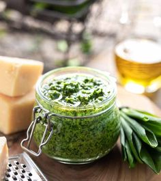 a glass jar filled with green pest next to sliced cheese