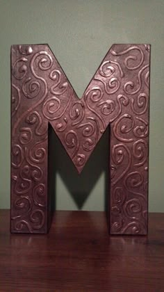 the letter m is made out of metal and sits on a wooden table next to a green wall