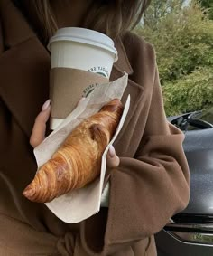 a woman holding a coffee cup and a croissant in her hand while standing next to a parked car