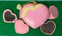 a pink heart shaped case with sunglasses, comb and mirror on green table top next to it