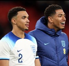 two young men standing next to each other on a soccer field, one is wearing a blue jacket and the other has a white shirt