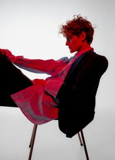 a young man sitting in a chair with his legs crossed and wearing a red shirt