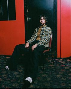 a man sitting on a chair in front of a red wall wearing a plaid shirt and black pants