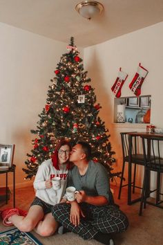 a man and woman are sitting in front of a christmas tree with their mugs