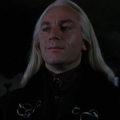 a man with long blonde hair wearing a black coat and chain around his neck is staring into the distance