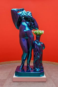 a blue statue is holding a vase with flowers in it on a pedestal next to a red wall