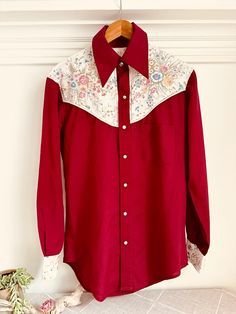 If there ever was an unspoken rule on the ranch, it was this; the tougher the person, the prettier the shirt.  And, my Uncle George and Aunt Nori wore a lot of them. This hand-sewn 1960s beauty features a pearl-snap front, oxblood base, and contrasting front and back wildflower-print yokes.  Truly a one-of-a-kind.  And truly a beauty. Measurements: Shoulders:  18 1/2" Sleeve:  24 5/8" Chest:  21 3/4" Overall Length:  33" Questions?  Please message us. Cheers! Vintage Cotton Tops For Country Events, Fitted Shirt For Western-themed Spring Events, Western Style Red Tops For Spring, Vintage Tops For Western-themed Events, Western Red Tops For Spring, Vintage Shirt For Rodeo In Fall, Vintage Shirt For Western-themed Fall Events, Vintage Cotton Tops For Rodeo, Spring Western Red Tops