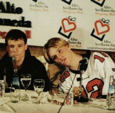 two young men sitting at a table with wine glasses in front of them and one man holding a microphone up to his ear