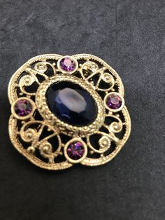 1928 Jewelry Company Victorian Brooch Blue Oval Faceted Glass Center, Purple Rhinestone Sets Filigree Style Quatrefoil Gold Finish Setting Victorian Revival Jewelry Set 1928 Jewelry Company Pressed Back Gold Tone Filigree Settings, Findings Vintage from 1970's-80's Size: 1 3/4 x 1 3/4 inches Condition: Very good; slight finish loss on pin back. Will be shipped First Class Mail in a gift box Blue Oval Brooch Jewelry, Oval Blue Brooch Jewelry, Blue Oval Brooch For Gifts, Oval Blue Brooch, Blue Oval Brooch, Elegant Oval Blue Brooches, Elegant Blue Oval Brooches, Vintage Sapphire Brooch Jewelry, Oval Brooch For Vintage Events