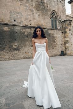 a woman standing in front of a brick building wearing a white dress and holding a bouquet