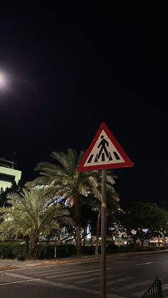 a pedestrian crossing sign on the side of a road at night with palm trees in the background