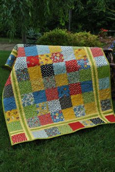 a patchwork quilt sitting on the grass in front of some trees and bushes,