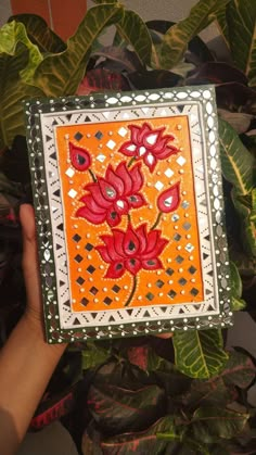 a hand holding an orange and white box with red flowers on it, surrounded by green leaves