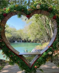 a heart shaped frame with flowers and greenery on the outside, in front of a lake