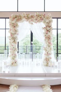 a white wedding arch decorated with flowers and candles in front of a large glass window