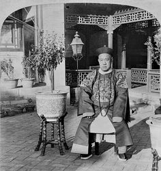 an old black and white photo of a man sitting on a chair in front of a building