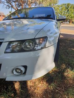 the front end of a white car parked on grass next to a tree and street