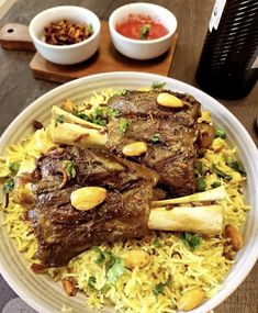 a white plate topped with meat and rice next to bowls of sauces on a wooden table