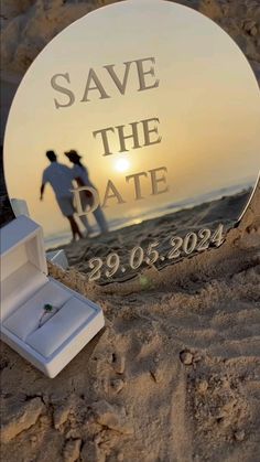 a round mirror with the words save the date and an engagement ring in it on sand