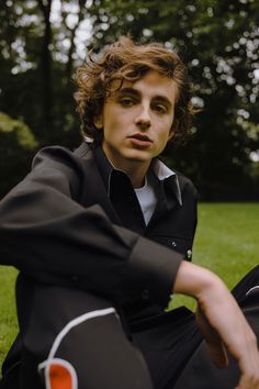 a young man with curly hair sitting on the grass in front of trees and looking at the camera