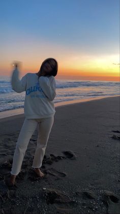 a woman standing on top of a sandy beach next to the ocean at sunset with her arms in the air