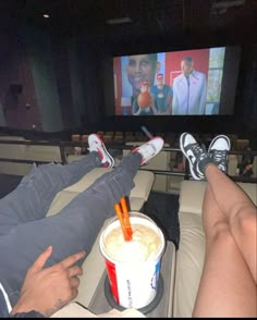 two people sitting in front of a movie screen with drinks on their laps and feet up